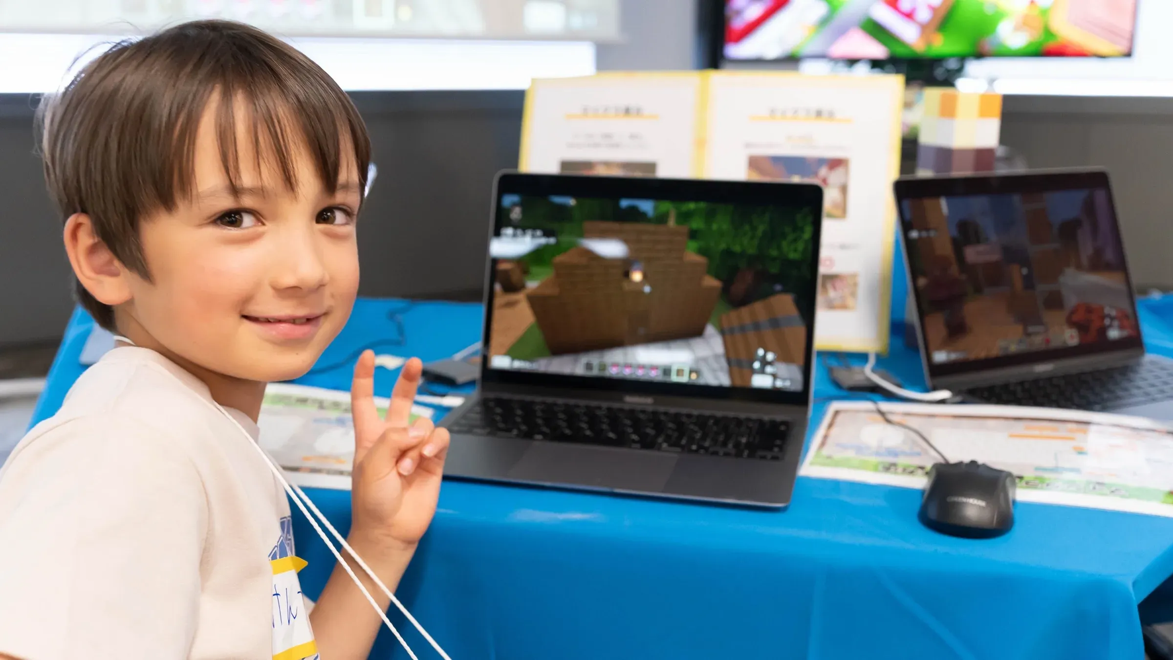 マインクラフトで建築した画面の前でピースサインをする子ども
