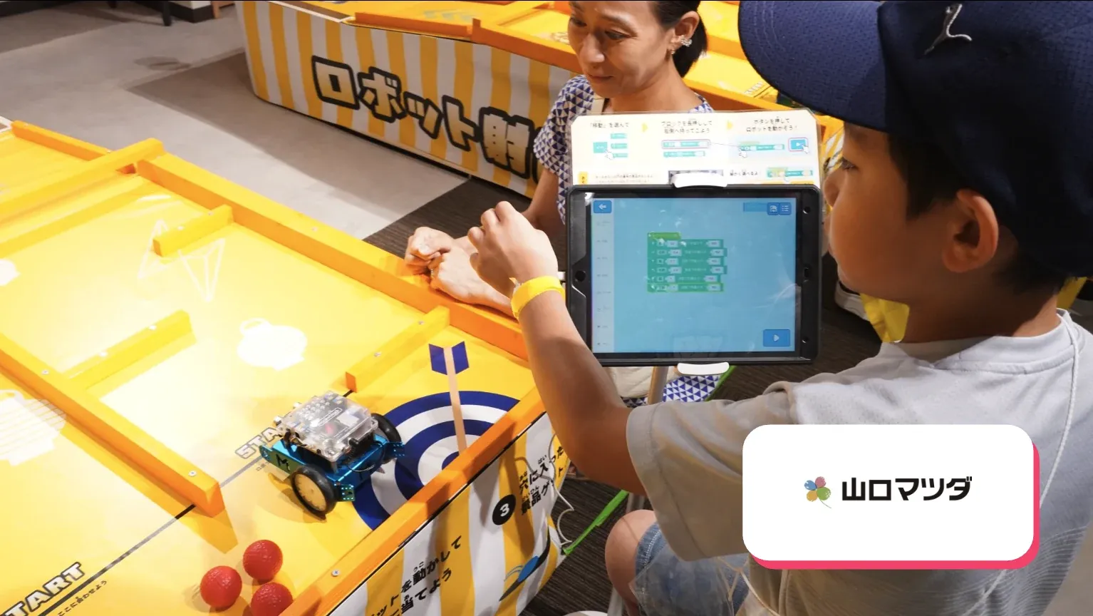タブレットで車型ロボットを操作する子どもと、山口マツダ株式会社のロゴ