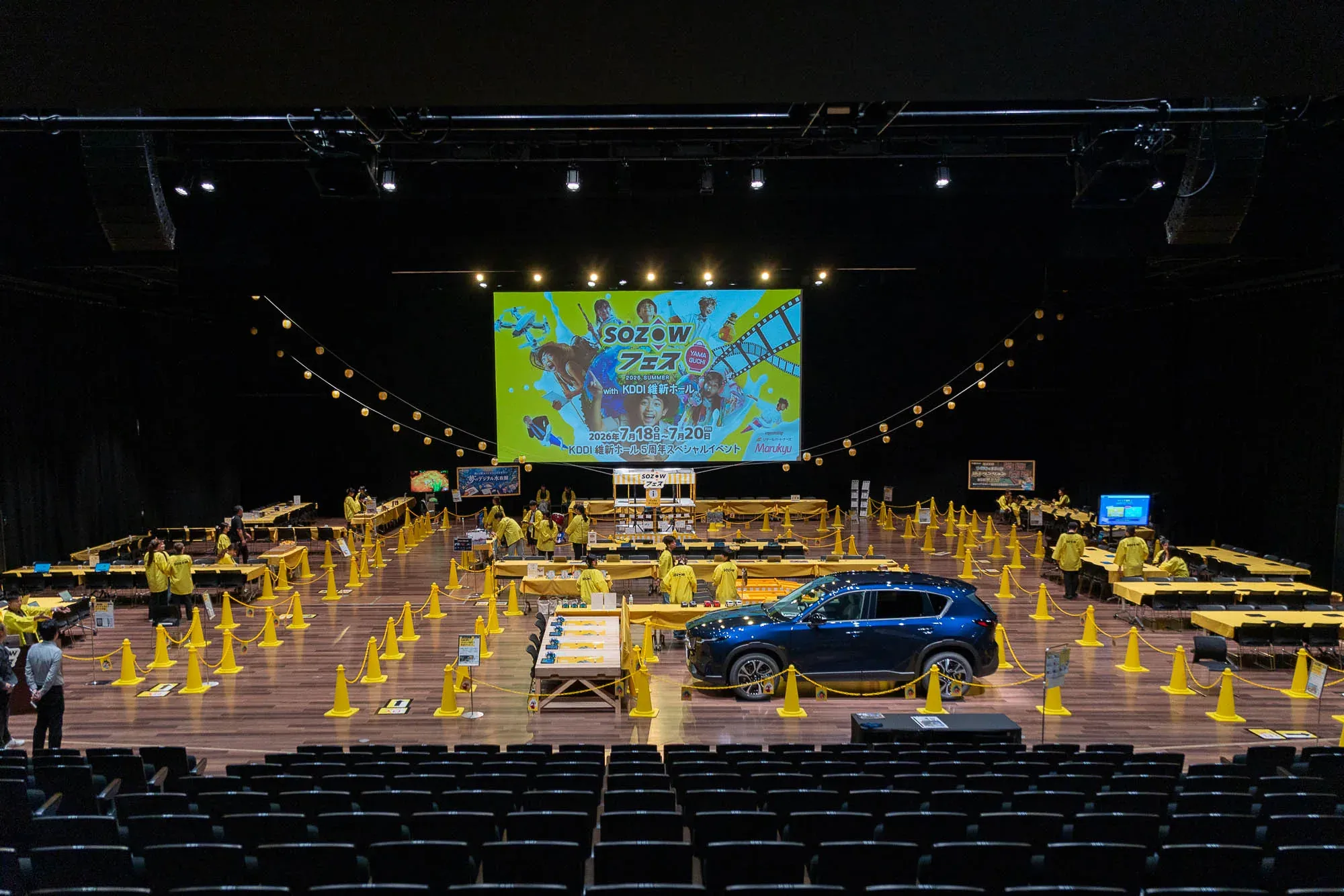 開場前のKDDI維新ホール メインホール。黄色いテーブルとコーンが並び、正面の大型スクリーンにSOZOWフェスのロゴが映っている