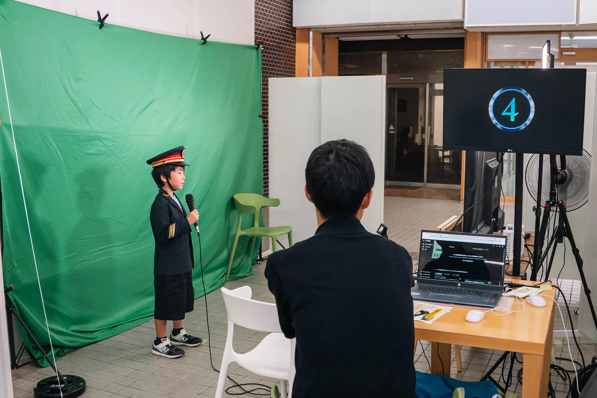 JR新山口駅南口で、駅員の制服を着てグリーンバックの前でアナウンスに挑戦する子ども