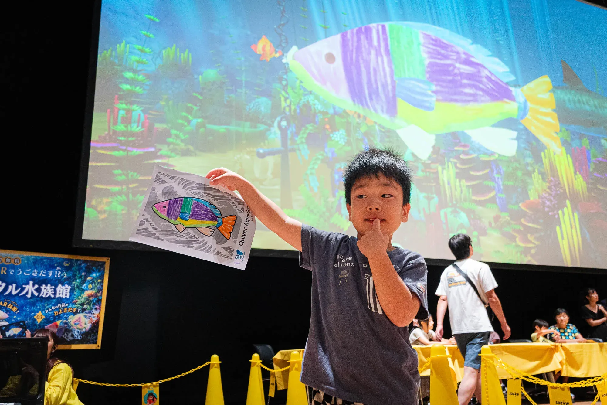 自分が描いた魚を掲げる子ども。背後の大画面には、その魚が泳ぐデジタル水族館が映っている