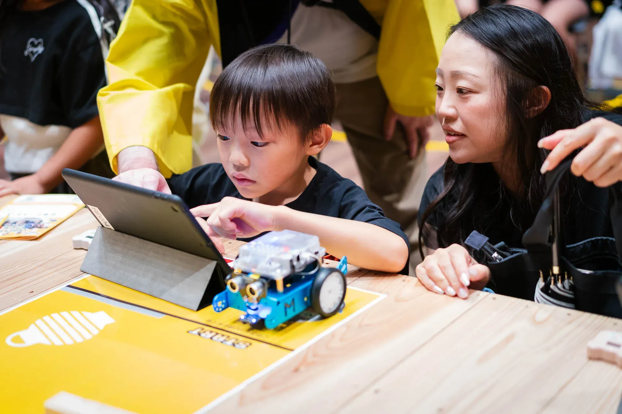 タブレットでロボットの動きをプログラミングする子どもと、それを見守る保護者