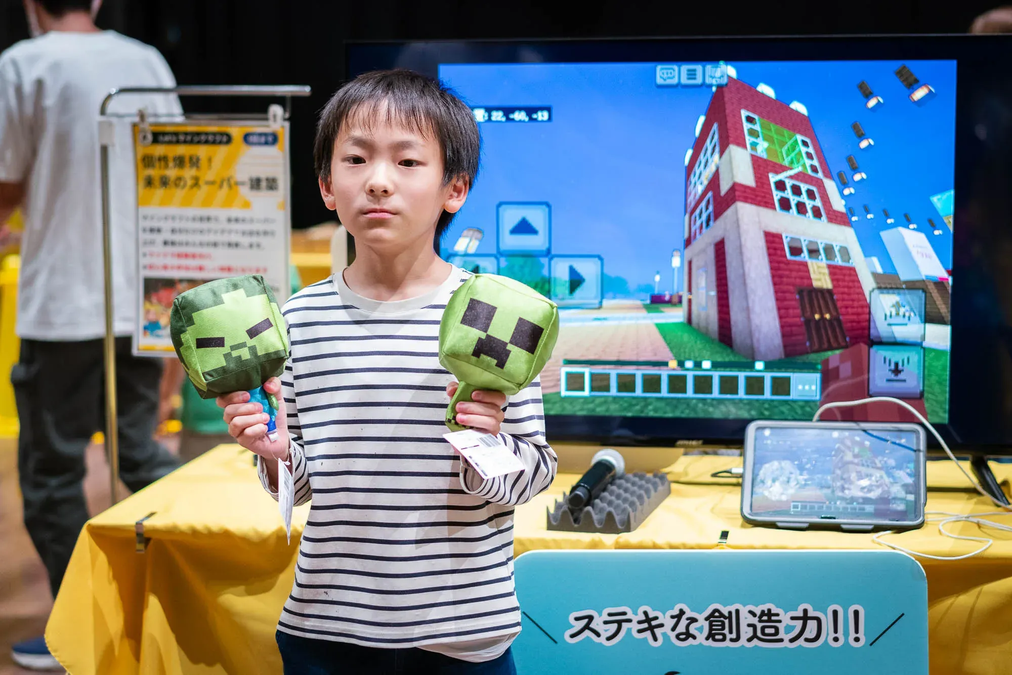 マインクラフトで建築した「未来のスーパー」を大型モニターに映し、その前でぬいぐるみを持って立つ子ども