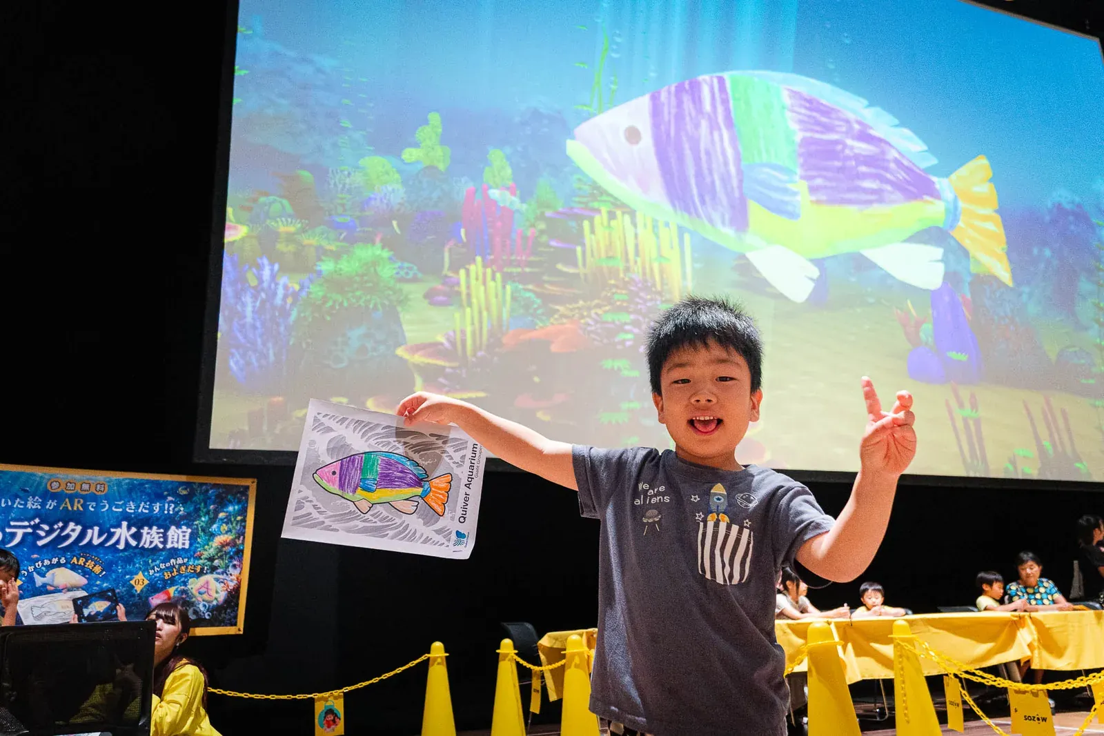 自分が描いた魚の絵を掲げてピースサインをする男の子。背後の大画面ではその魚が泳いでいる
