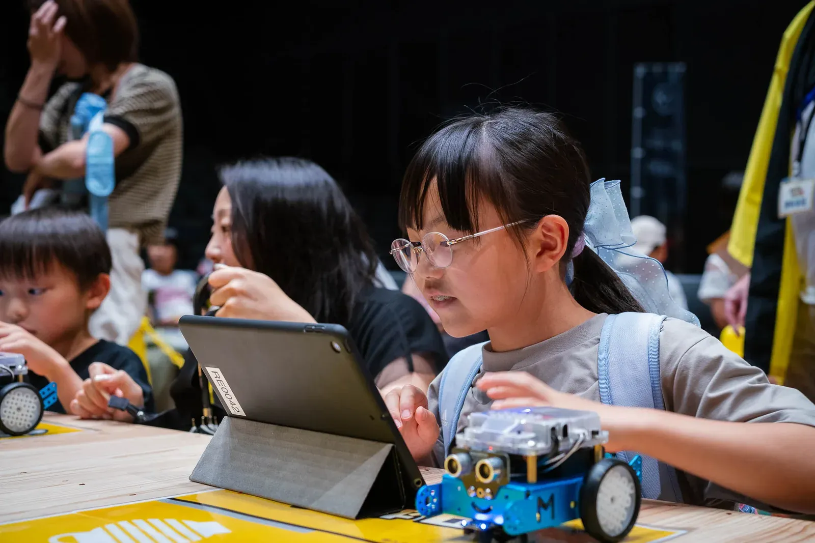 タブレットでロボットの動きをプログラミングする女の子と、机の上の車型ロボット